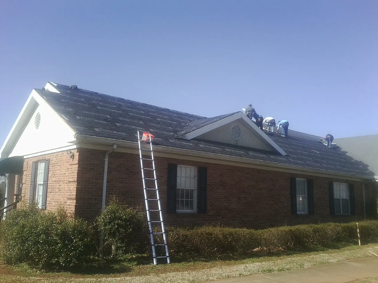 Crew tearing shingles off a brick ranch roof with a ladder set — Emergency Roof Repair work by our crew in Mantee