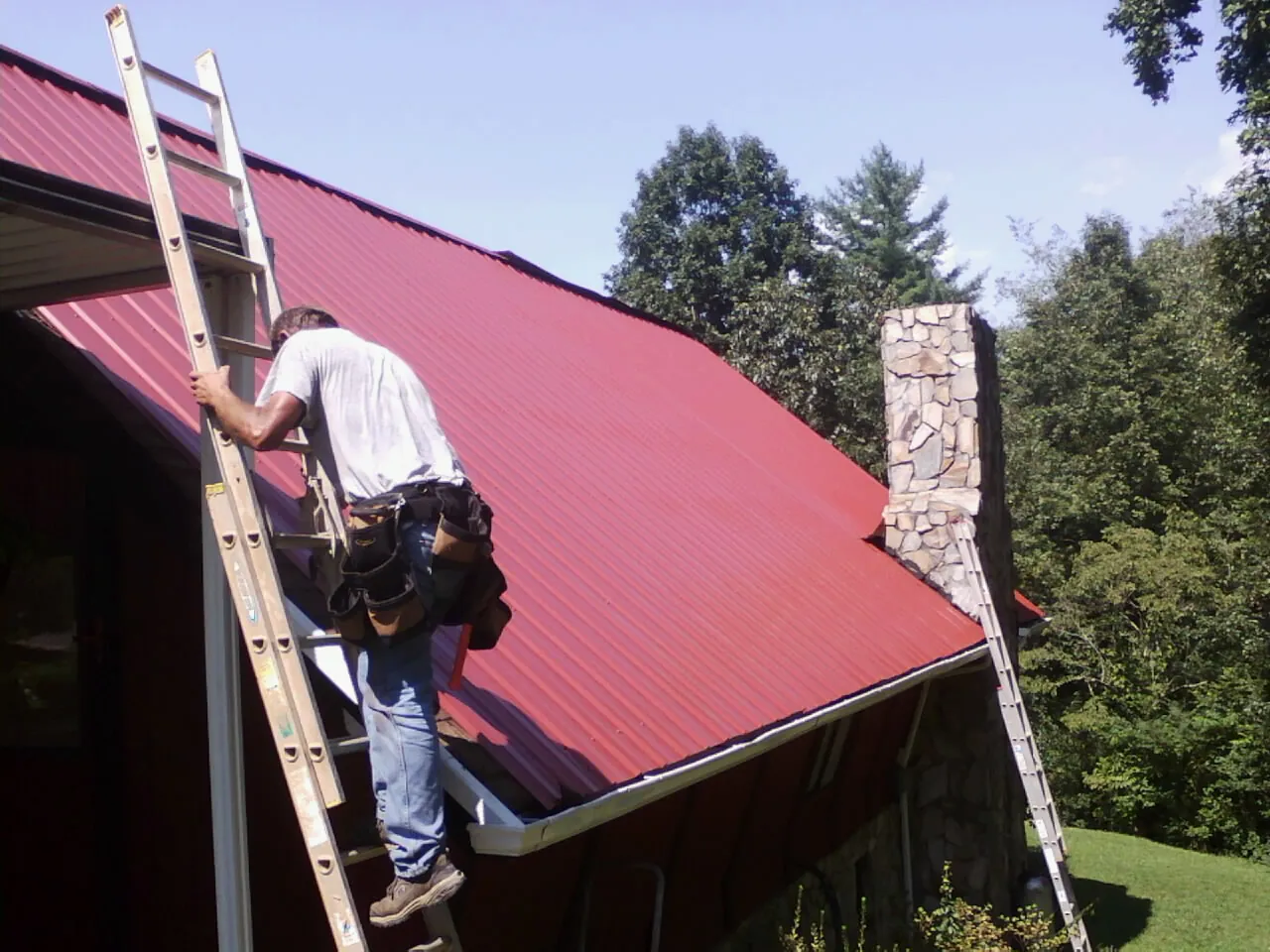 Roofer on a ladder beside a red metal roof and stone chimney — Soffit & Fascia Repair by Roofing Brothers in Mantee